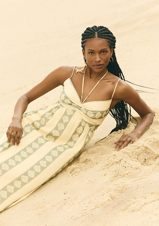 Modelo negra de rança deitada sobre areia da praia usando vestido longo com estampa de conchas