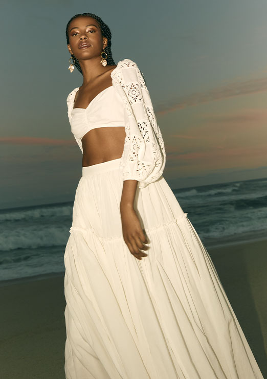 Modelo negra de trança na praia no entardecer usando conjunto branco de top com manga longa e bufante e saia solta da nova coleção de alto verão 25 Calmaria.