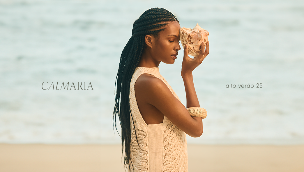 Modelo negra usando vestido de tricot na cor bege e pulseira de madeira no braço direito segurando uma concha perto do rosto, com o fundo da paisagem de mar e areia da praia. O banner tem o texto Calmaria alto verão 25.