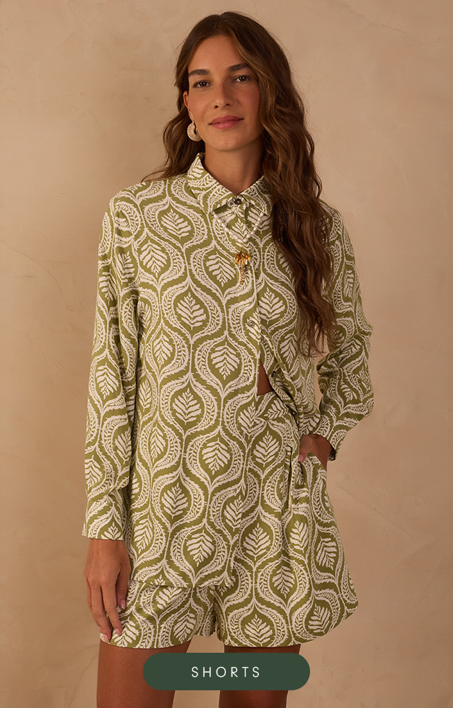 Modelo de pele clara e cabelos ruivos ondulados posa em um fundo bege texturizada, vestindo um conjunto estampado com lurex de saia longa soltinha com cadarço para amarração e barra marias e na parte de cima blusa de mangas longas com punhos ajustados com elástico, decote reto e comprimento cropped. O acessório no pescoço é um colar lenço com flor em metal dourado. A imagem possui um botão na cor vinho com texto "Até R$499".