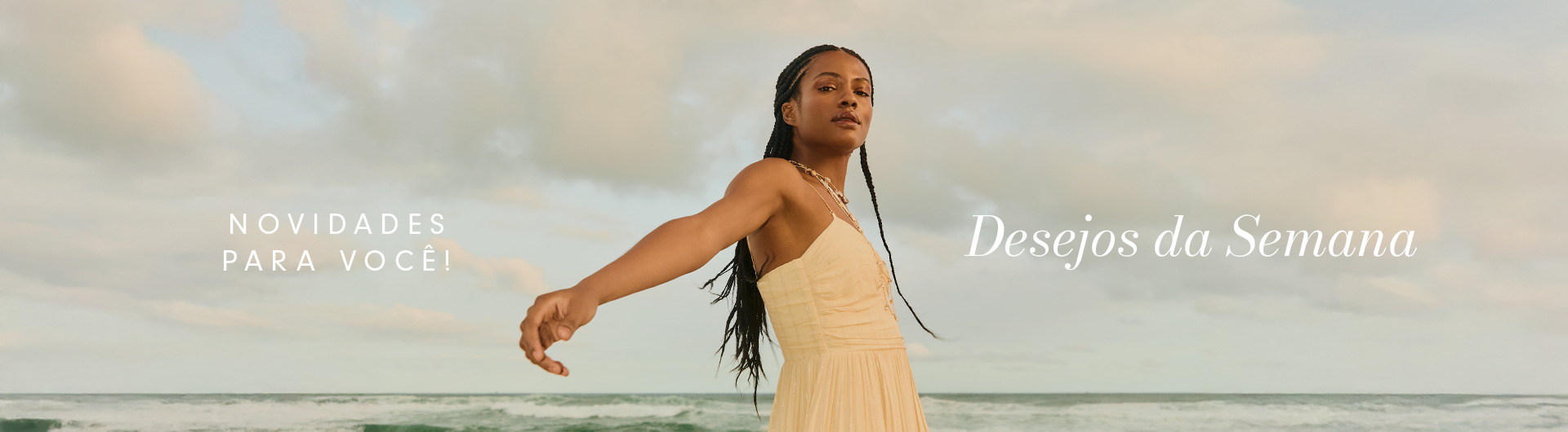 Imagem com fundo de céu e mar, ao entardecer com nuvens claras. No centro da imagem, uma modelo negra, usando vestido amarelo de alcinha da linha festa da coleção Calmaria. No texto, ao lado esquerdo: "Novidades para você!" e ao lado direito da modelo "Desejos da semana" na cor branco.