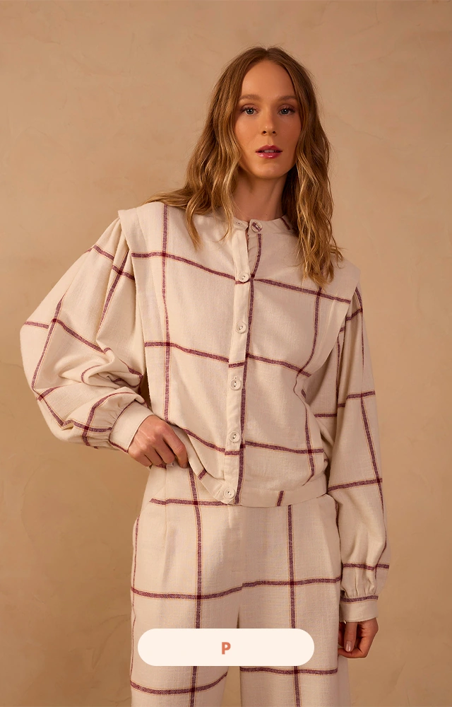 A modelo usa conjunto feminino xadrez off-white com detalhes em vinho, composto por jaqueta de modelagem ampla com mangas volumosas e calça de caimento fluido.