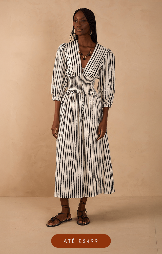 Modelo de pele clara e cabelos ruivos ondulados posa em um fundo bege texturizada, vestindo um conjunto estampado com lurex de saia longa soltinha com cadarço para amarração e barra marias e na parte de cima blusa de mangas longas com punhos ajustados com elástico, decote reto e comprimento cropped. O acessório no pescoço é um colar lenço com flor em metal dourado. A imagem possui um botão na cor vinho com texto "Até R$499".