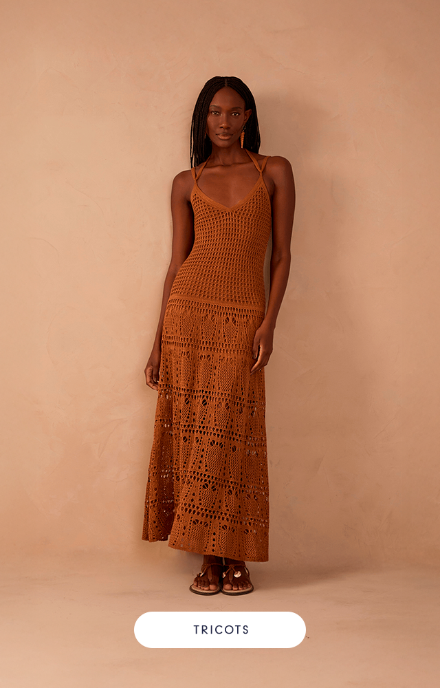 Modelo feminina contra fundo bege, usando macacão branco de tecido leve. A peça é longa, com pernas largas e alças finas cruzadas no colo. Nos pés, rasteira marrom.