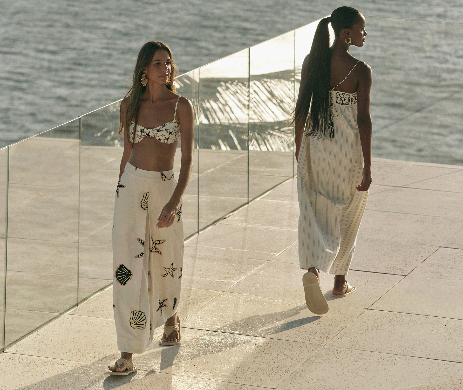 Duas modelos, um loira e uma preta desfilando em fachada de casa de praia, ambas usam peças da nova coleção de verão 25 da Dress To, nas cores offwhite, com detalhe de listras e detalhes de concha e estrela do mar.