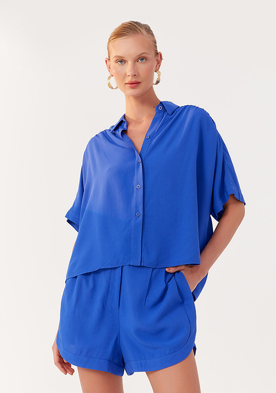 Modelo em fundo claro, usando conjunto azul de camisa de botão e short curto, coleção Praia à Vista