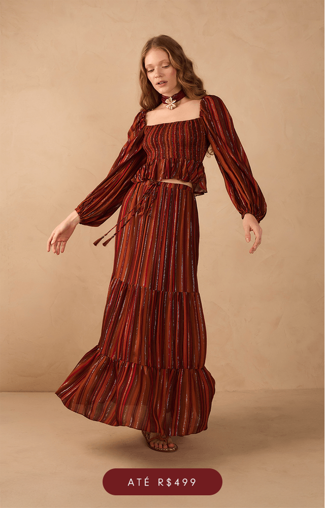 "Modelo com cabelos longos e ondulados posa em ambiente de fundo bege texturizado, usando conjunto de calça e colete em tecido com listras verticais nas cores vinho e terracota, com estampas sutis em tom mostarda. A calça é de modelagem ampla e reta, enquanto o colete possui botões frontais e bolsos decorativos. Nos pés, a modelo usa sapatos baixos em tom caramelo. Abaixo, botão com a palavra 'Calças'. 