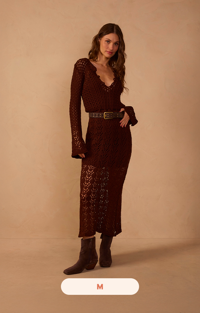 A modelo usa vestido midi em tricot rendado marrom, com decote em V, mangas longas e cintura marcada por cinto. Elegância, conforto e sofisticação em uma única peça.