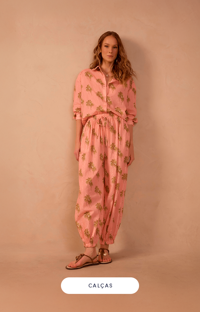 Modelo loira com cabelos curtos, usando blusa estilo regata soltinha de tricot dourado, calça pantalona branca com faixa para amarração na cintura e rasteira na cor bronze. A imagem tem fundo bege com botão verde escrito "calças" na cor branca.