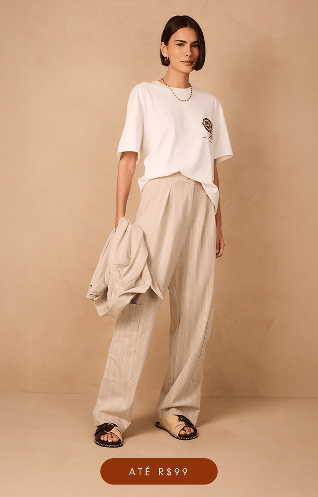 Modelo de pele clara e cabelos ruivos ondulados posa contra uma parede bege texturizada, vestindo um conjunto de t-shirt silkada e saia listrada branca e vermelha. Além de usar um colar com pingente de coração. A imagem possui um botão na cor vinho com texto "Até R$199".