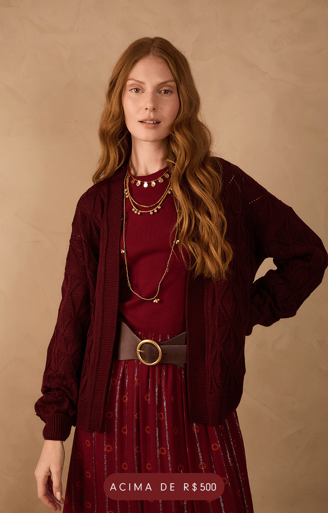 Mulher com cabelos longos e ondulados vestindo conjunto boho vinho com estampa discreta e brilho sutil. A blusa possui mangas amplas e decote profundo com amarração frontal. A saia longa é fluida, com cintura alta e babado na barra. Ela usa sandálias rasteiras e colares dourados. Fundo bege neutro. Botão com a palavra 'SAIAS' em destaque na parte inferior da imagem.