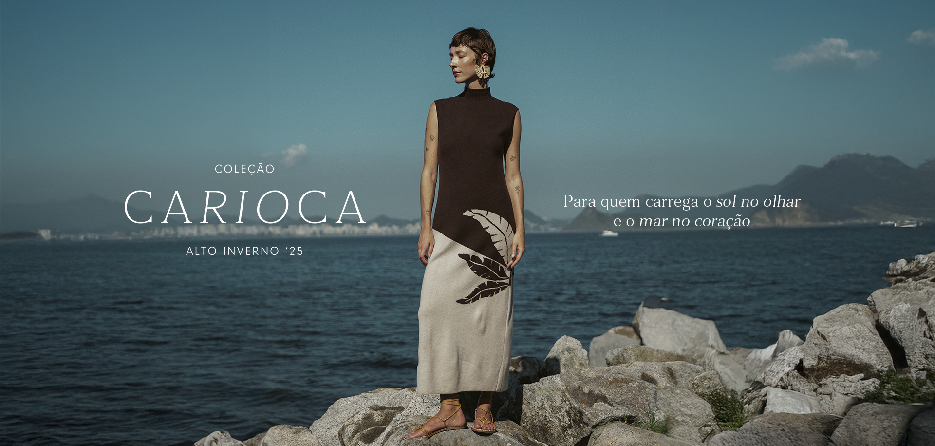 Banner da coleção Carioca da Dress To, Alto Inverno 2025. Modelo usa vestido sem mangas marrom e bege com estampa de folhas, posando sobre pedras à beira-mar, com o Pão de Açúcar ao fundo.