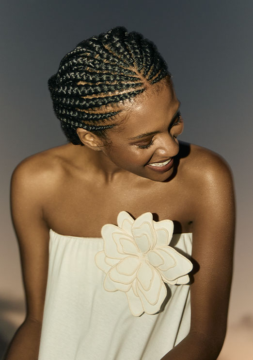 Modelo morena usando cabelo de trança e vestido de tomara que caia na cor branco com um detalhe de flor no busto.