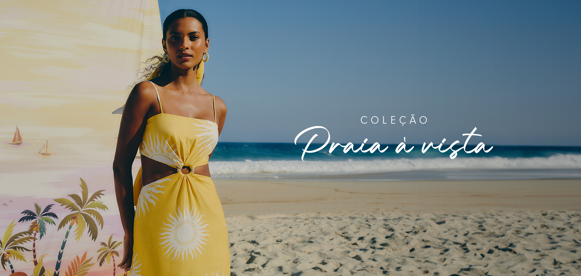 Modelo na praia utilizando vestido com recortes laterais, argola frontal e alça fina, na cor amarela com estampa de sol da coleção praia à vista.