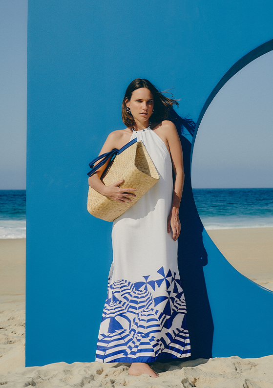 Modelo na praia usando vestido com estampa na barra e bolsa de palha, coleção praia à vista