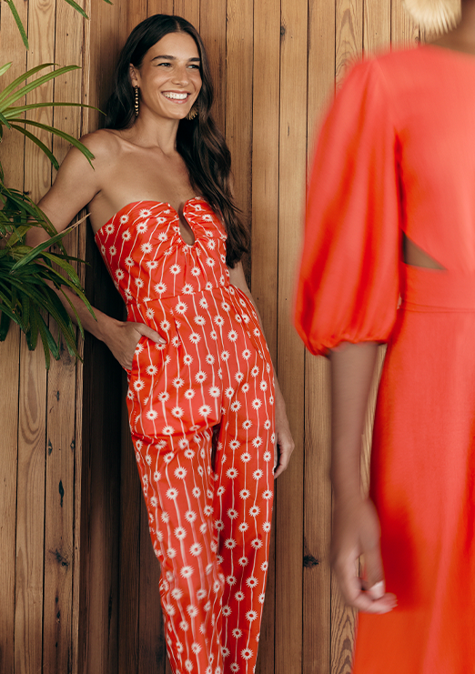 Modelo morena usando macacão vermelho com flores brancas, encostada na parede de madeira.