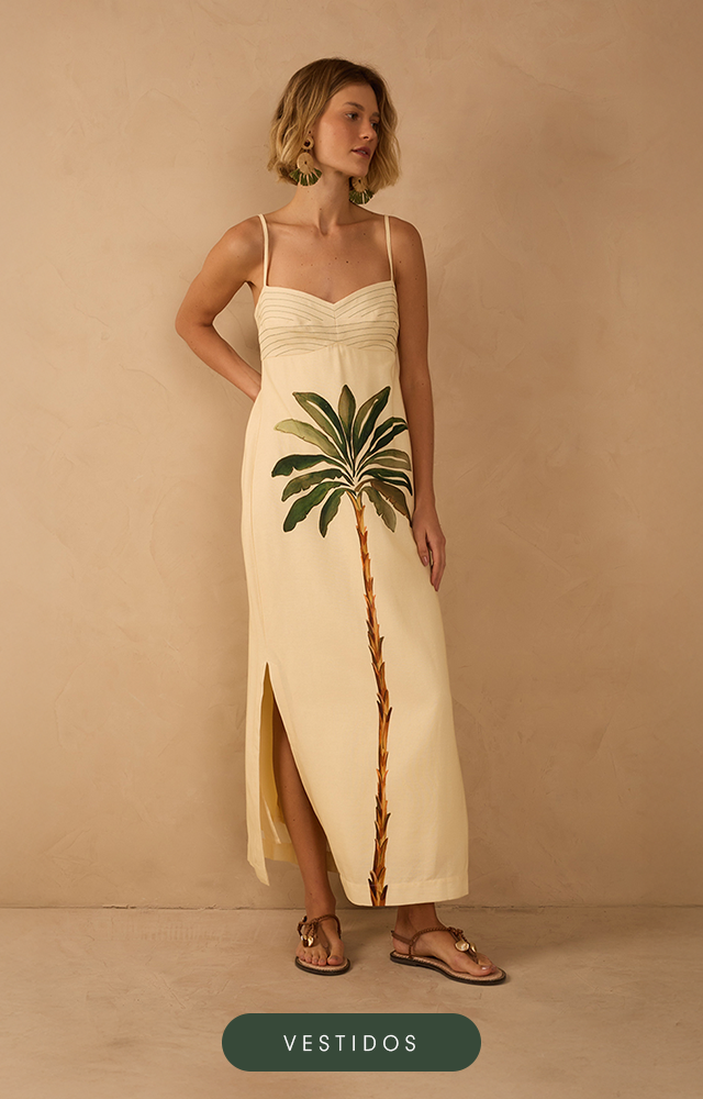 Modelo de pele clara e cabelos ruivos ondulados posa contra uma parede bege texturizada, vestindo um conjunto de t-shirt silkada e saia listrada branca e vermelha. Além de usar um colar com pingente de coração. A imagem possui um botão na cor vinho com texto "Até R$199".