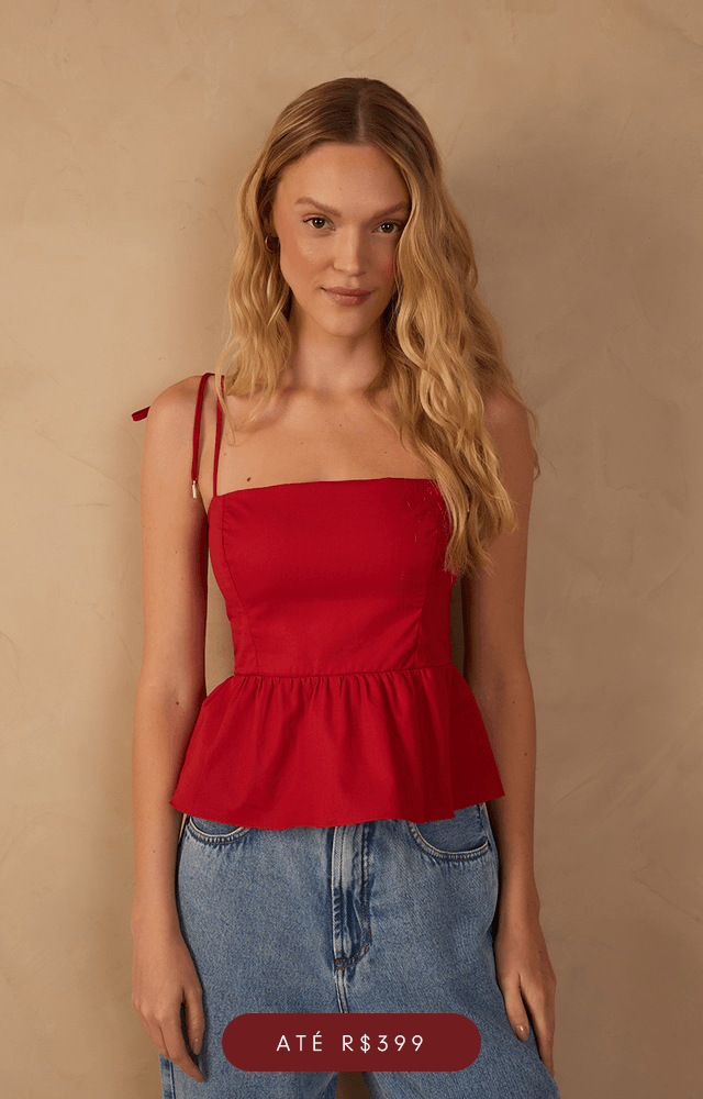 Modelo de cabelos ruivos ondulados posa em frente a parede bege texturizada, vestindo blusa cropped de manga longa e ampla em tecido leve, na cor vinho com estampas pequenas em laranja e branco. A peça tem decote em V profundo com amarração central e detalhe de pingente dourado. A modelo também usa colares dourados em camadas. Na parte inferior da imagem, botão com a palavra 'Blusas'.