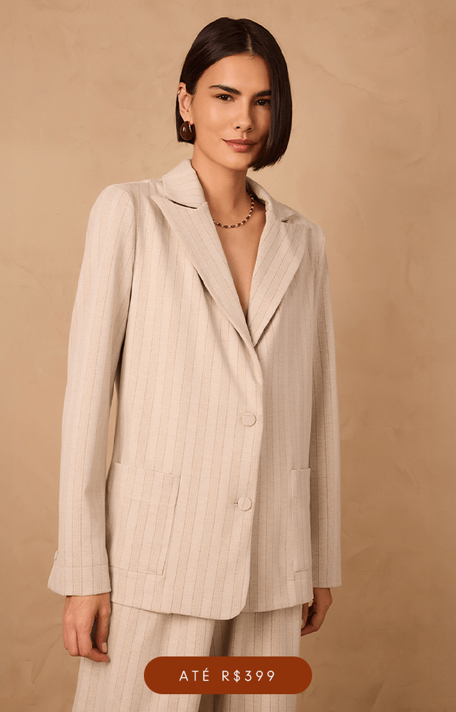 Modelo de pele clara e cabelos ruivos ondulados posa em um fundo bege texturizada, vestindo um conjunto estampado com lurex de saia longa soltinha com cadarço para amarração e barra marias e na parte de cima blusa de mangas longas com punhos ajustados com elástico, decote reto e comprimento cropped. O acessório no pescoço é um colar lenço com flor em metal dourado. A imagem possui um botão na cor vinho com texto "Até R$499".